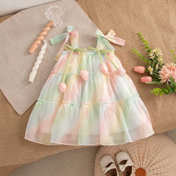 A pastel rainbow tiered baby dress with bow straps and tulip embellishments, styled in a flat-lay arrangement on a beige fabric background with decorative props.